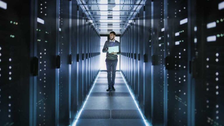 IT Technician Works on Laptop in Big Data Center full of Rack Servers. He Runs Diagnostics and Maintenance, Sets System Up.